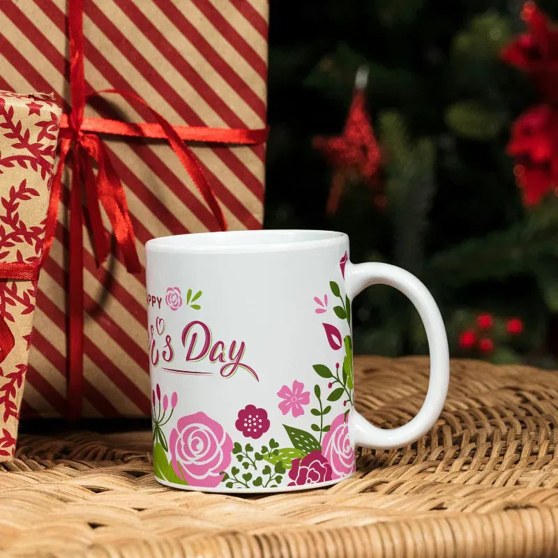 White mug with floral designs and 'Happy Mother's Day' text, placed on a woven surface with gift boxes and flowers in the background.