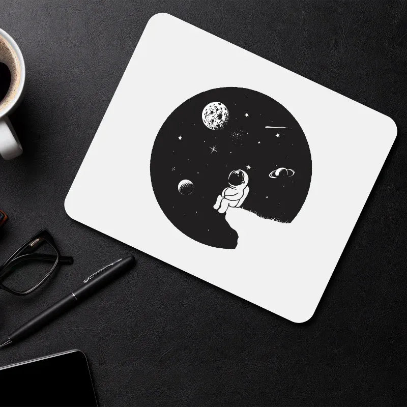 White mouse pad with astronaut design on a black surface with a cup, pen, and glasses.