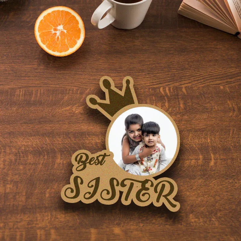 Decorative sign with photo of two children and 'Best Sister' text on a wooden surface with an orange and mug.