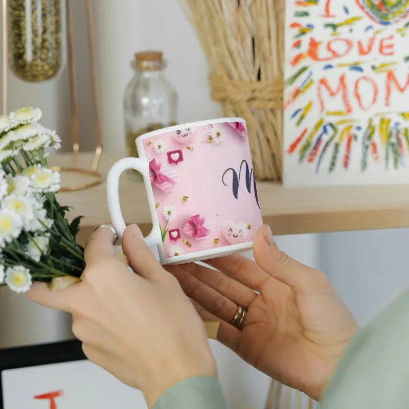 Person holding a mug with floral design and 'Mom' text, surrounded by flowers and a 'I love Mom' sign.