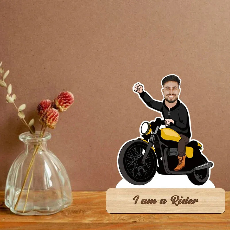 Figurine of a man on a motorcycle with 'I am a Rider' text on a wooden base, next to a vase with flowers.