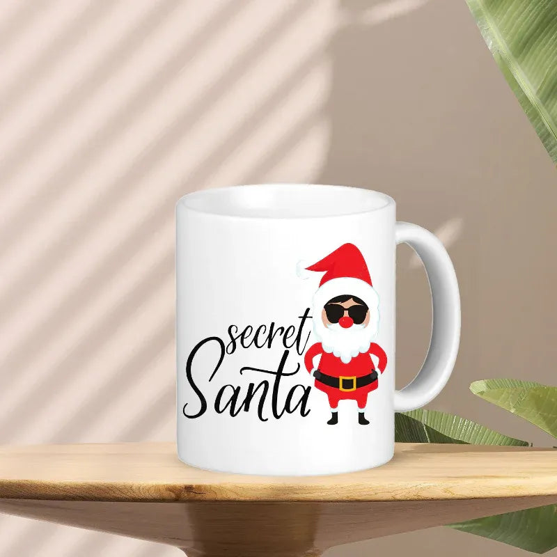 White mug with a cartoon Santa Claus design and 'secret Santa' text on a wooden surface.