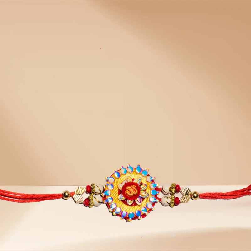 A traditional Indian Rakhi bracelet with a red cord, featuring a central gold-toned medallion decorated with red, blue, and white stones, and small elephant-shaped beads on either side.