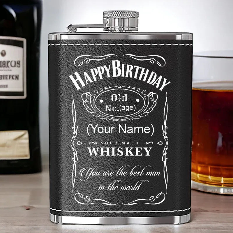 Personalized whiskey flask with 'Happy Birthday' message on a wooden surface with a glass of whiskey.