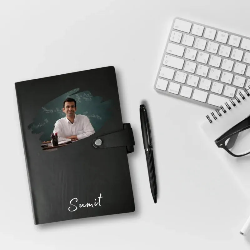 Black notebook with a photo of a man on the cover, placed on a white surface with a keyboard and pen.