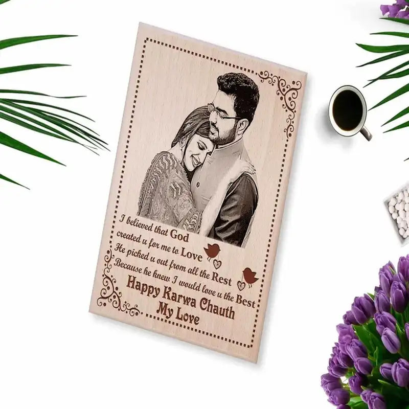 A personalized wooden engraved Karwa Chauth photo frame featuring a photo of a couple and a romantic quote, styled on a white surface with decorative green leaves, a cup of coffee, and purple flowers.