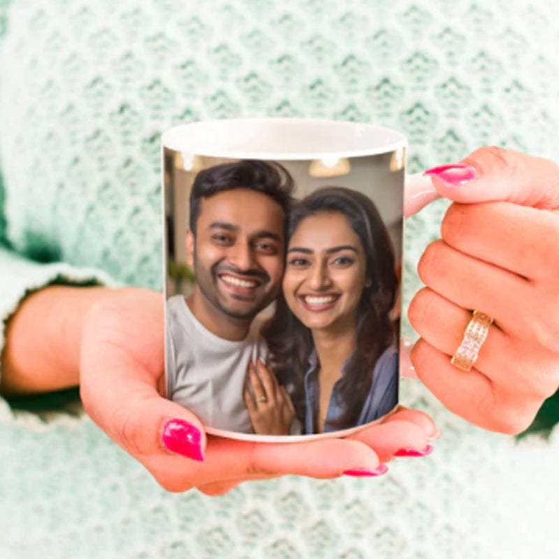 A customized ceramic picture mug with a photo of a couple holding a camera, being held by a person with pink nails.