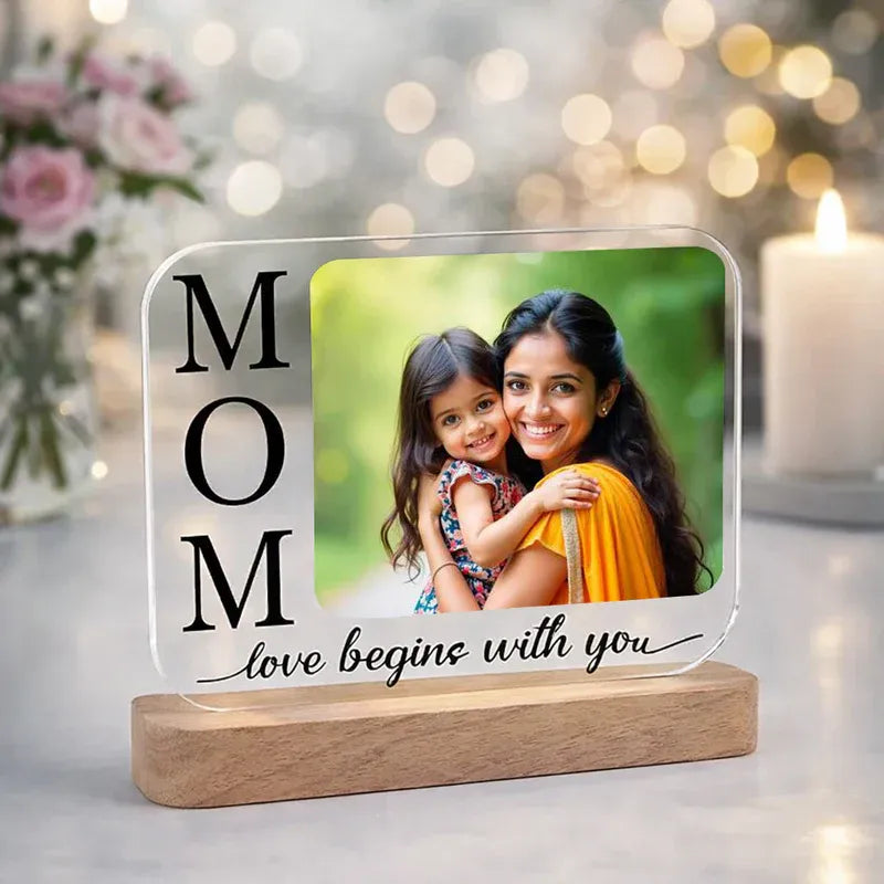 Decorative acrylic photo frame with a photo of a woman and child, text 'MOM love begins with you', on a wooden stand with a blurred background.