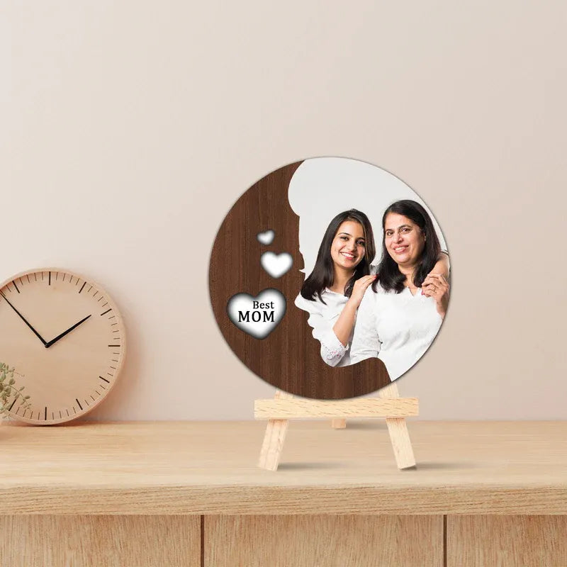 Round Mother's Day photo frame with a cutout of two women and 'Best Mom' text on a stand, with a clock in the background.