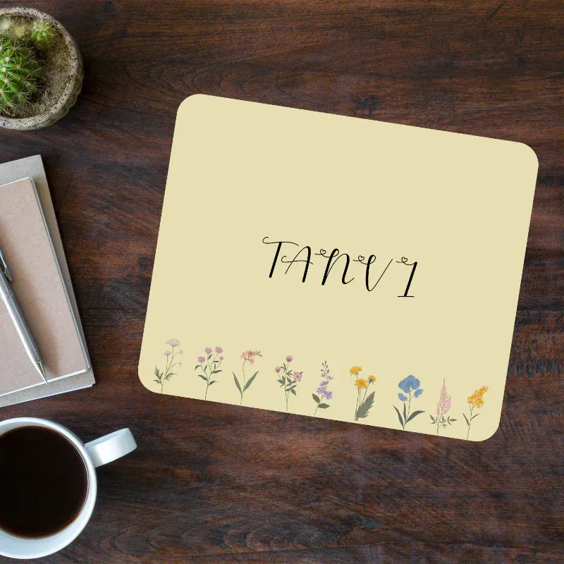 Beige placemat with floral designs and name personalized on a wooden table with a cup of coffee and small plant.