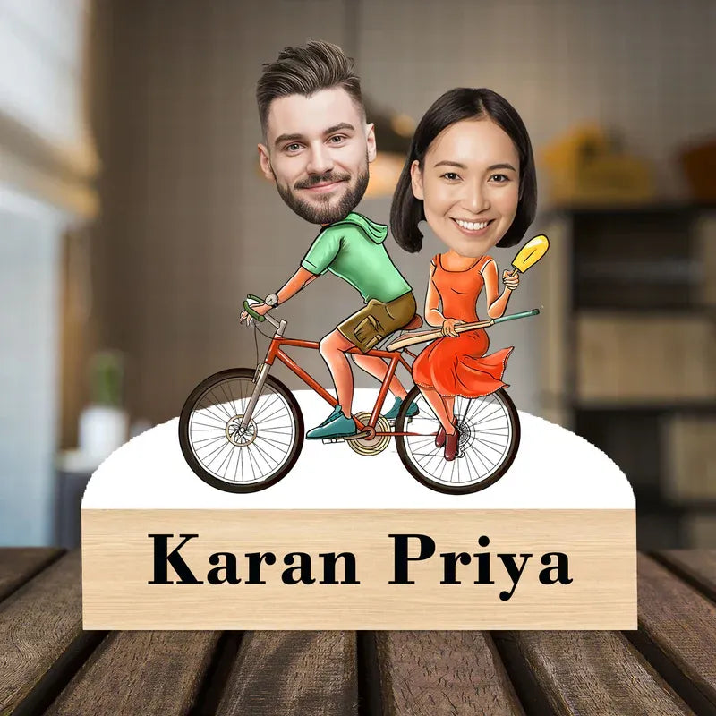 Cartoon depiction of a man and woman on a bicycle with 'Karan Priya' below, set against a blurred indoor background.