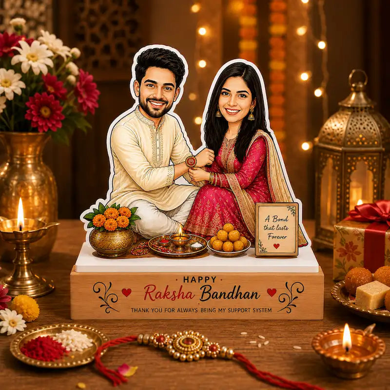 A decorative Raksha Bandhan display featuring a die-cut cardboard cutout of a smiling couple seated together on a wooden base. The base is printed with 'HAPPY Raksha Bandhan' and includes small decorative items such as marigold flowers, sweets, and a small sign reading 'A Bond that lasts Forever.