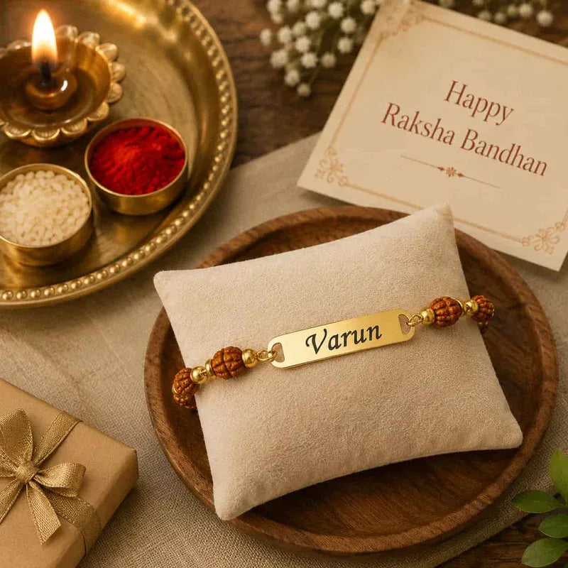 A personalized Raksha Bandhan bracelet with a gold nameplate engraved 'Varun' and brown bead accents, displayed on a beige cushion on a wooden tray. The scene is decorated with a lit diya, a card reading 'Happy Raksha Bandhan', and a gift box.