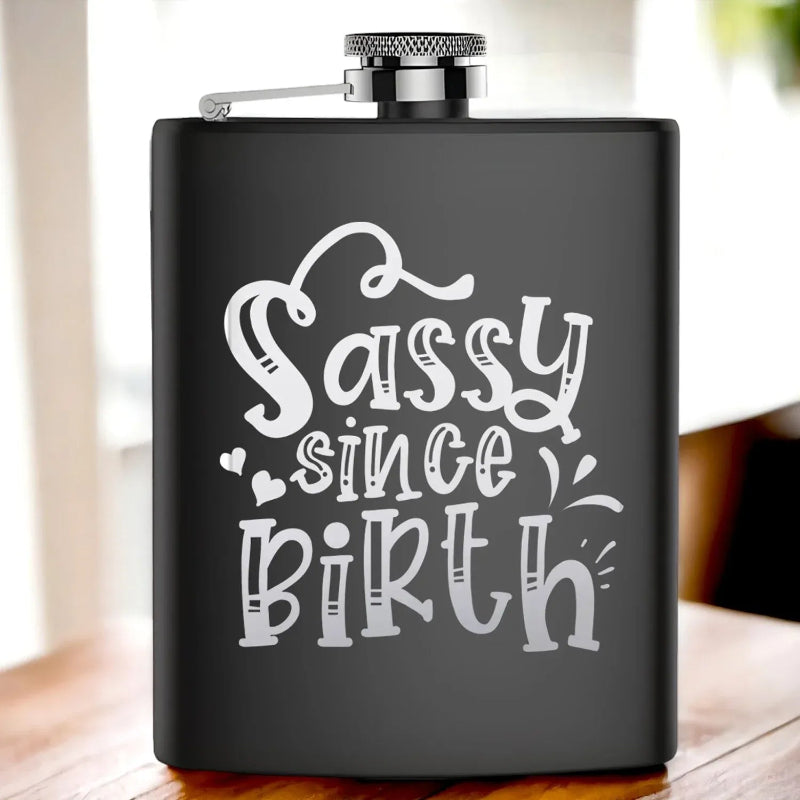 Black flask with 'Sassy since birth' text on a wooden surface