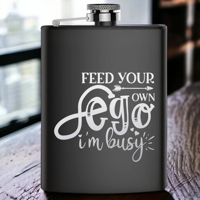 Black flask with white text 'Feed your own ego, I'm busy' on a wooden surface.