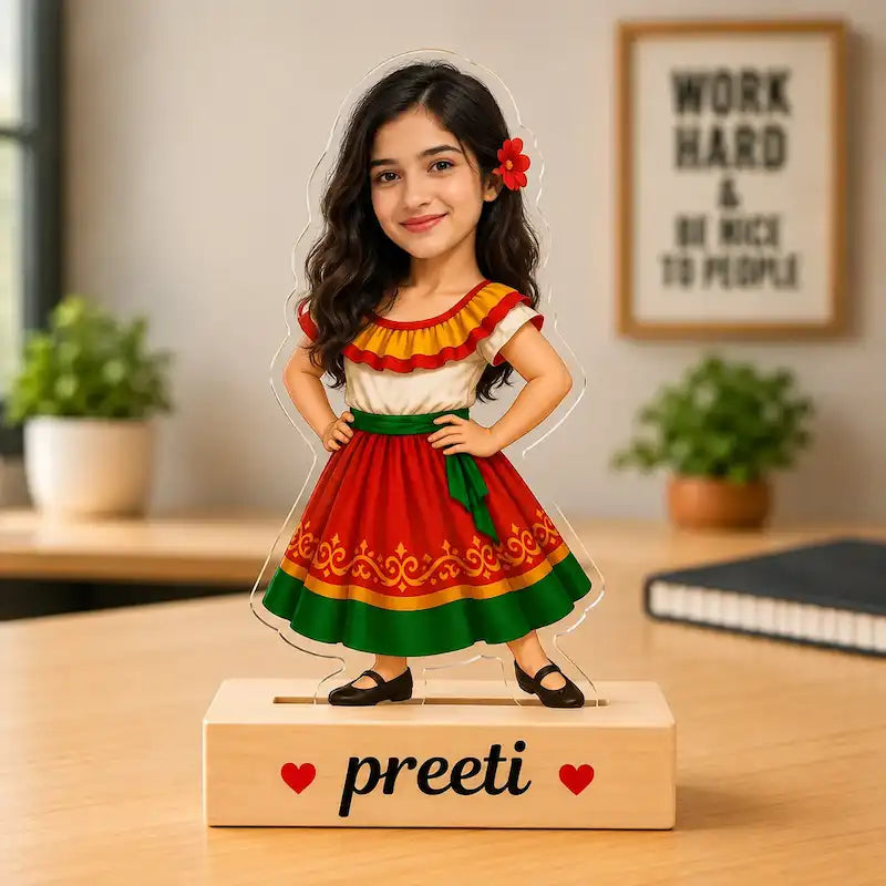 A personalized acrylic standee of a young girl with long dark hair, wearing a red, yellow, and green traditional-style dress, standing on a wooden base engraved with the name 'preeti' and small red hearts. The stand is placed on a wooden desk with a blurred office background containing plants and a framed sign.