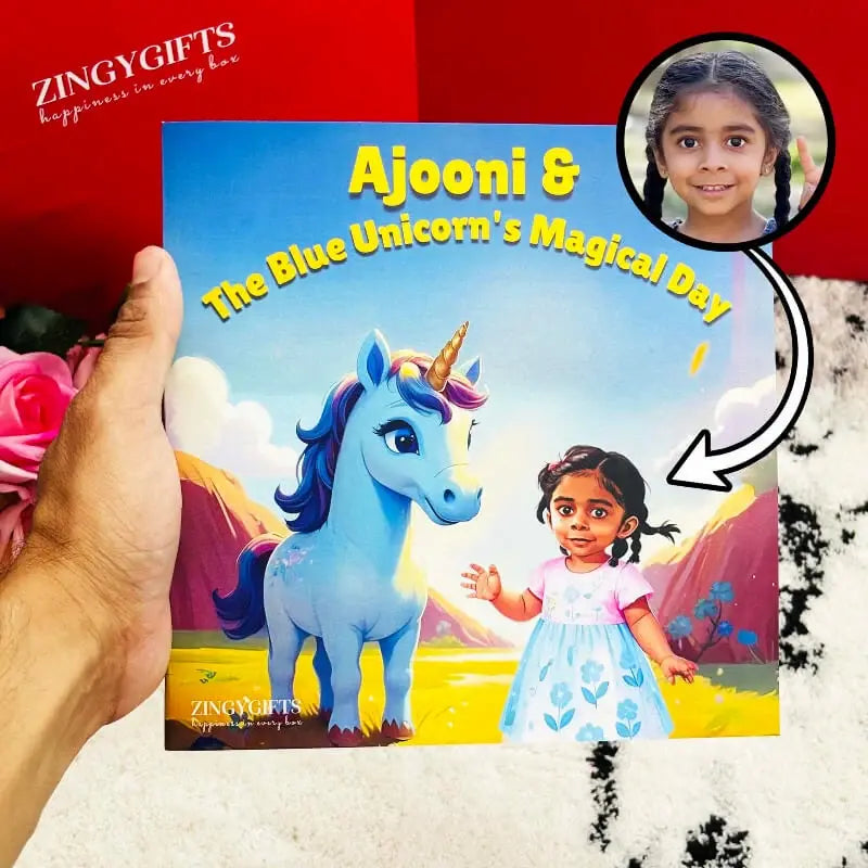 A hand holds a personalized children's picture book titled 'Ajooni & The Blue Unicorn's Magical Day' against a red and white textured background. The cover features a blue unicorn and a young girl.