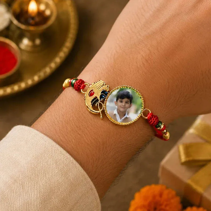 A gold-plated personalized Rakhi bracelet with a photo of a boy, a small Ganesha charm, and red and gold beaded accents, shown worn on a person's wrist.
