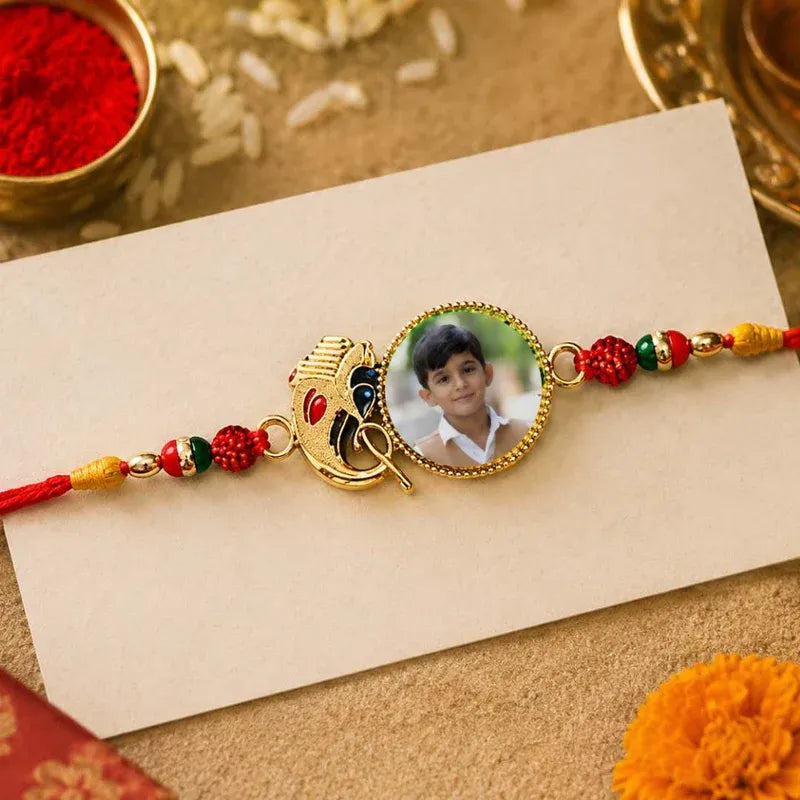 A personalized gold-plated baby boy's Rakhi bracelet displayed on a cream-colored card. The bracelet features a central oval photo of a young boy, a gold Peacock charm, and red, green, and yellow decorative beads, set against a festive background with rice and red powder.