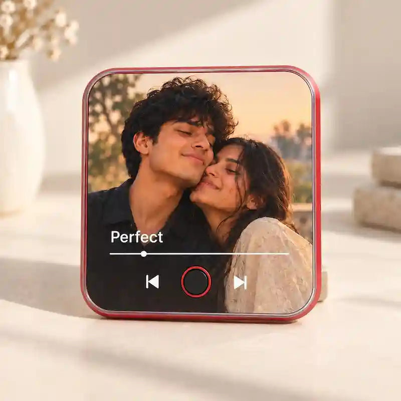 A personalized music fridge magnet device showing a custom photo and the song 'Until I Found You' by Stephen Sanchez & Em Behhold. The background is a cozy, softly blurred living room with a lit candle, a cup of tea, and a bookshelf.