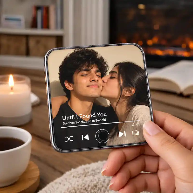 A personalized music fridge magnet device showing a custom photo and the song 'Until I Found You' by Stephen Sanchez & Em Behhold. The background is a cozy, softly blurred living room with a lit candle, a cup of tea, and a bookshelf.