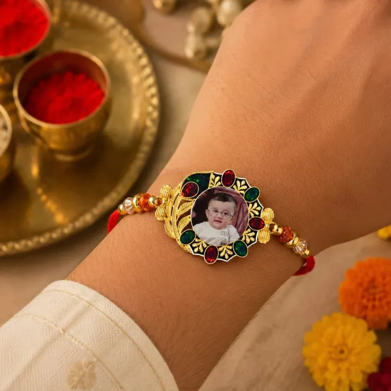 A close-up of a person's wrist wearing a decorative gold-plated Rakhi bracelet with red and green enamel details, a central photo insert, and red beads. A traditional brass tray with red powder and flowers is blurred in the background.