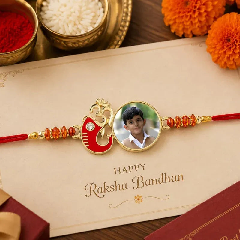 A personalized Rakhi with a circular photo of a young boy, a red and gold Om symbol, and red thread, displayed on a 'Happy Raksha Bandhan' card with festive props.