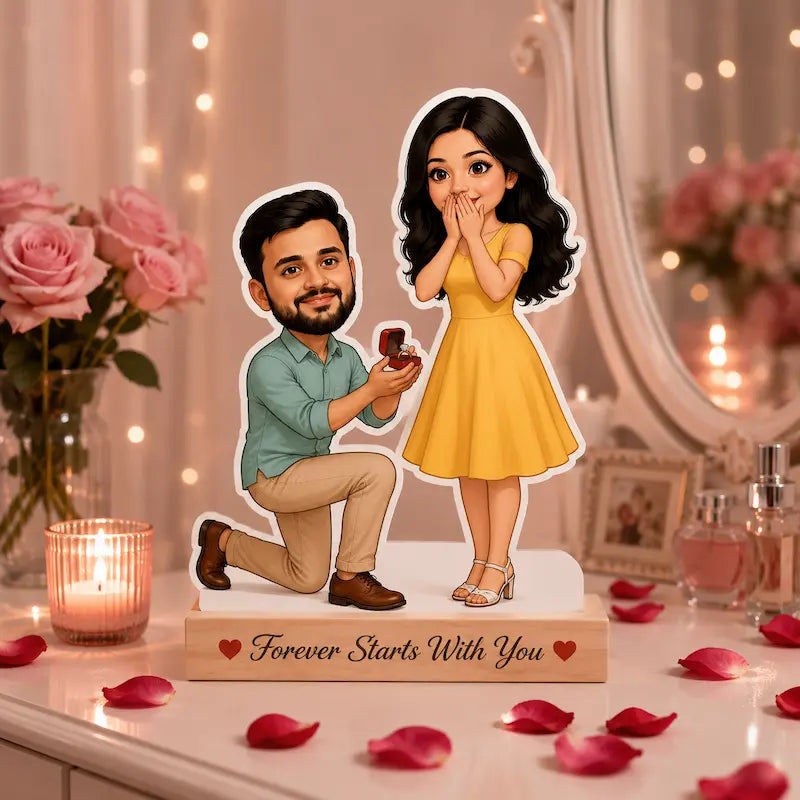 A personalized cartoon couple figurine displayed on a decorated table. The cutout shows a man kneeling and proposing with a ring box, and a woman in a yellow dress, mounted on a wooden base that reads 'Forever Starts With You'.