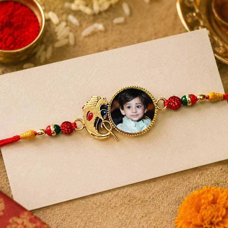 A personalized Rakhi featuring a circular gold-toned medallion with a photo of a young boy, flanked by a peacock charm and red, green, and gold decorative beads, laid on a cream-colored card over a textured beige surface with festive props.