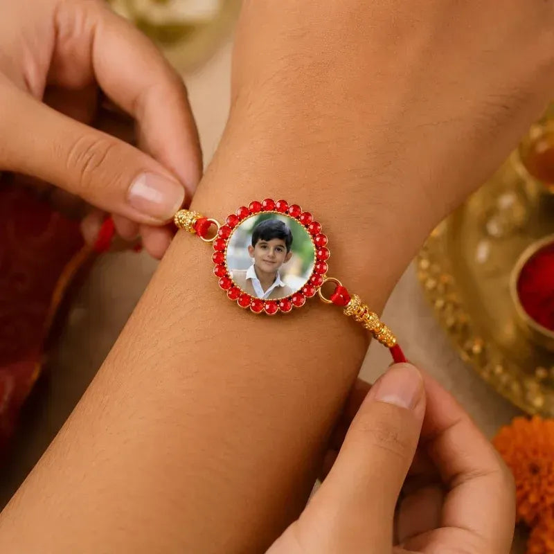 A close-up of a person's wrist wearing a personalized Rakhi bracelet. The bracelet features a red cord, gold-toned decorative beads, and a central oval photo frame surrounded by red stones.