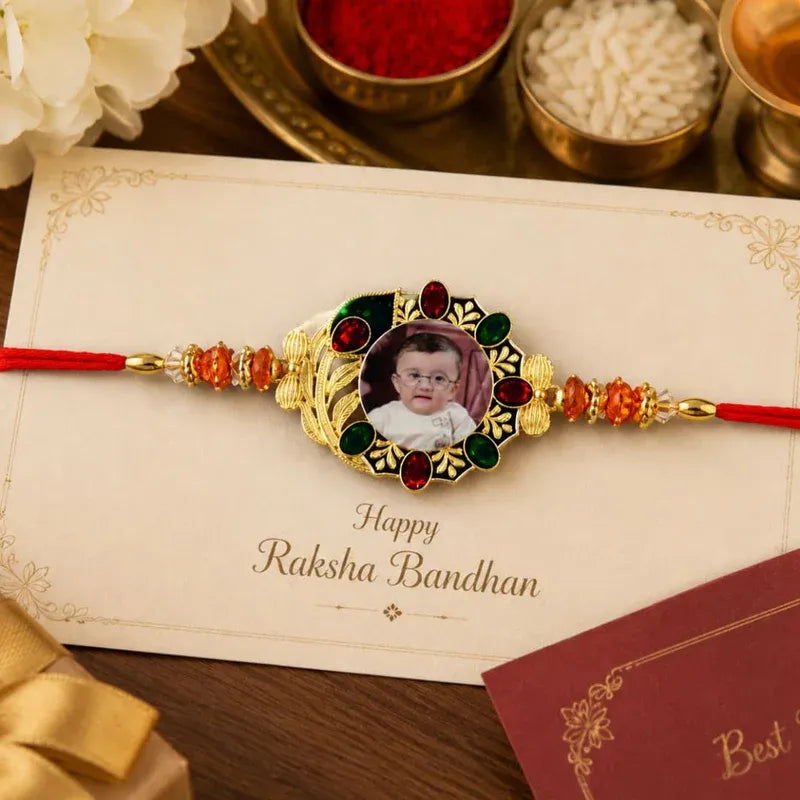 A personalized Rakhi with a photo of a baby, set on a cream-colored greeting card that reads 'Happy Raksha Bandhan'. The Rakhi features a gold-toned central frame with red and green stone accents and red cords on either side, styled on a wooden surface with festive decorations in the background.