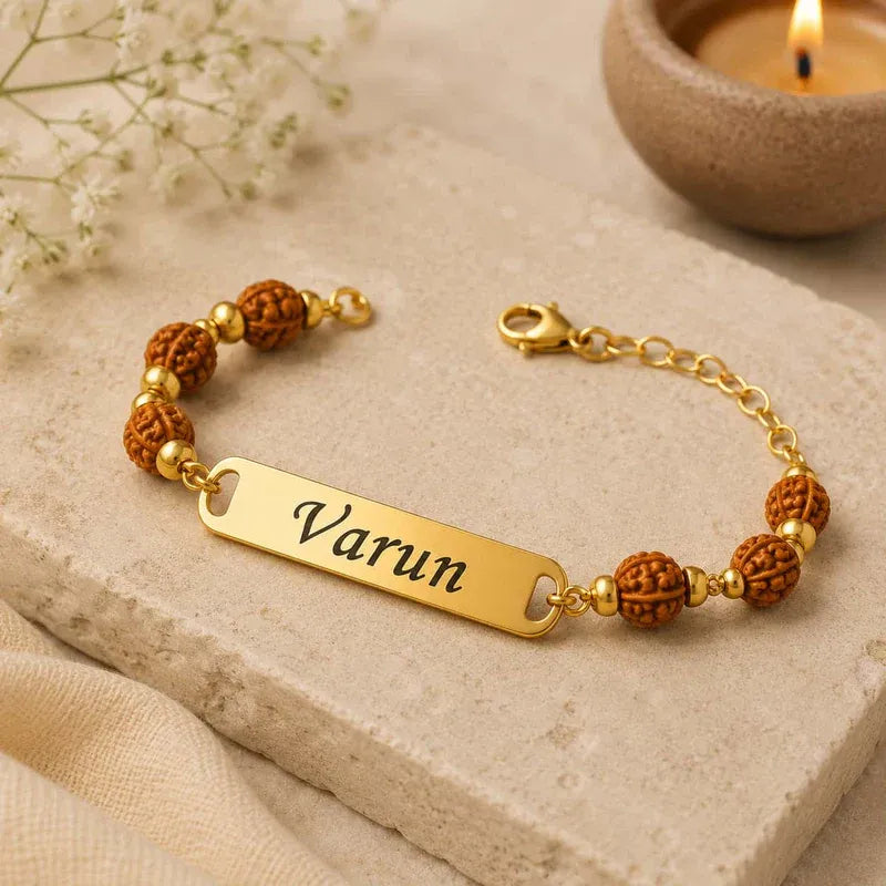 A personalized gold bracelet with a rectangular nameplate engraved 'Varun' and brown textured beads, styled on a beige stone slab with a lit candle in the background.
