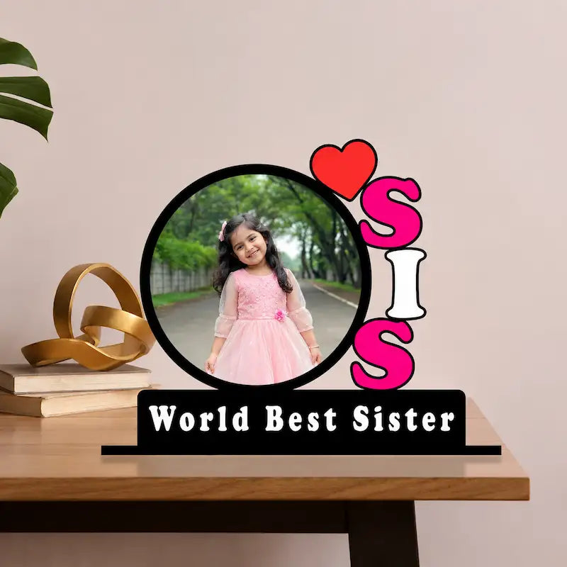 A personalized tabletop photo sign with a circular frame holding a young girl in a pink dress, surrounded by the word 'SIS' in bright pink, white, and red letters, with a black base reading 'World Best Sister.' The sign is styled on a wooden table against a beige wall, with decorative books and a gold ribbon beside it.