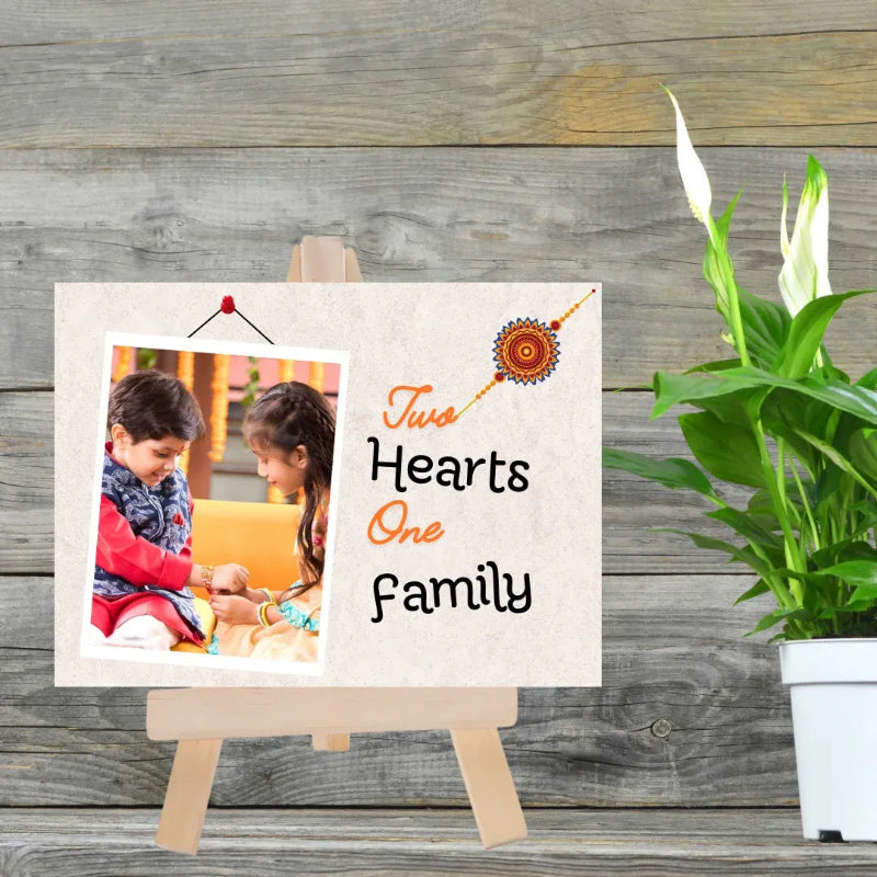 A personalized wooden photo plaque on a small easel, featuring a custom photo of two children and the text 'Two Hearts One Family' in black and orange lettering, styled on a rustic wood background next to a potted plant.