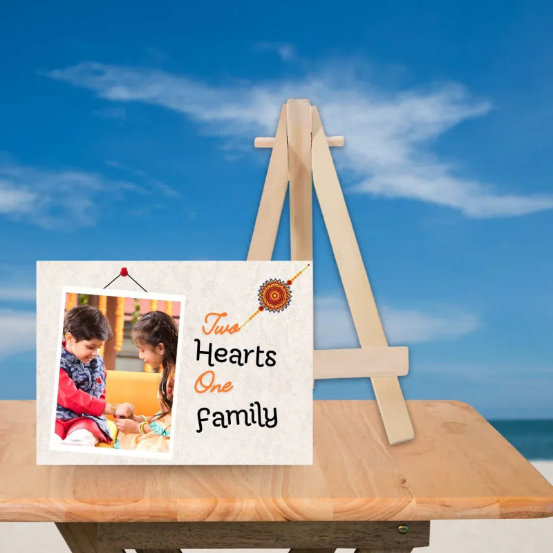 A personalized wooden picture frame displayed on a beach easel. The frame features a custom photo of two children on the left and the text 'Two Hearts One Family' with a small orange and yellow decorative emblem on the right, all set against a blue sky background.