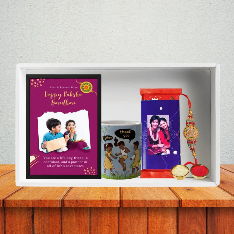 A personalized Rakhi gift set displayed inside a white wooden tray on a wooden table against a textured wall. The set includes a purple greeting card with a photo of a couple, a colorful illustrated mug, a purple wrapped chocolate with a photo, and a red and gold Rakhi thread.