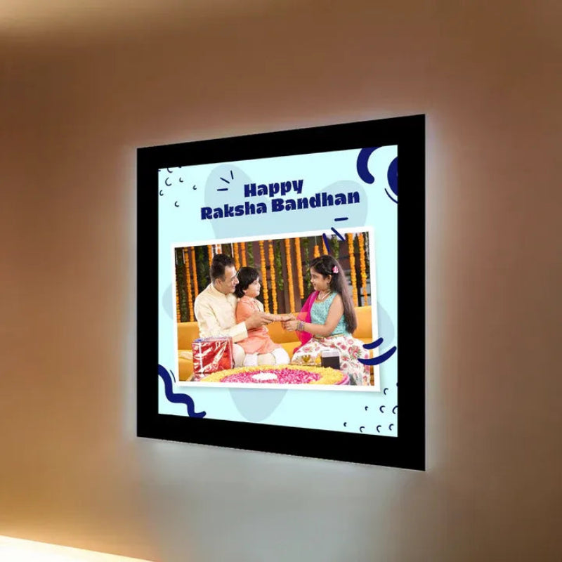 A square LED light box display mounted on a beige wall, showcasing a custom photo with 'Happy Raksha Bandhan' text and decorative blue graphics.