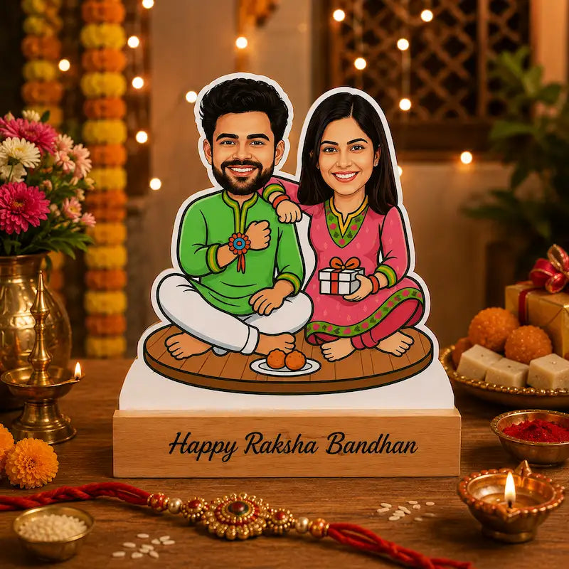 A custom cartoon couple cutout displayed on a wooden base with the text 'Happy Raksha Bandhan' on a decorated wooden table. The cutout depicts a smiling man in a green kurta and white pants and a woman in a pink kurta holding a gift box.