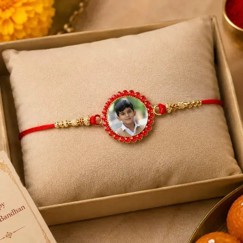A red and gold Rakhi bracelet with a custom photo insert of a young boy, displayed on a beige cushion inside a gift box.