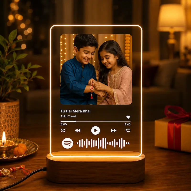 A glowing rectangular smart speaker or music player on a wooden base sits on a table in a warmly lit living room. The screen shows a photo of a boy and girl and a Spotify-style playback interface with the text 'Tu Hai Mera Bhai' and 'Aastha Tensei.