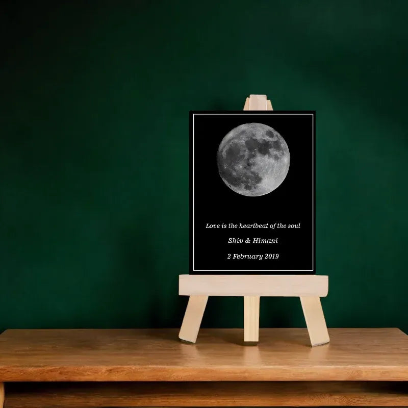 Moon image with personalized text on a black frame, placed on a wooden stand against a green wall.
