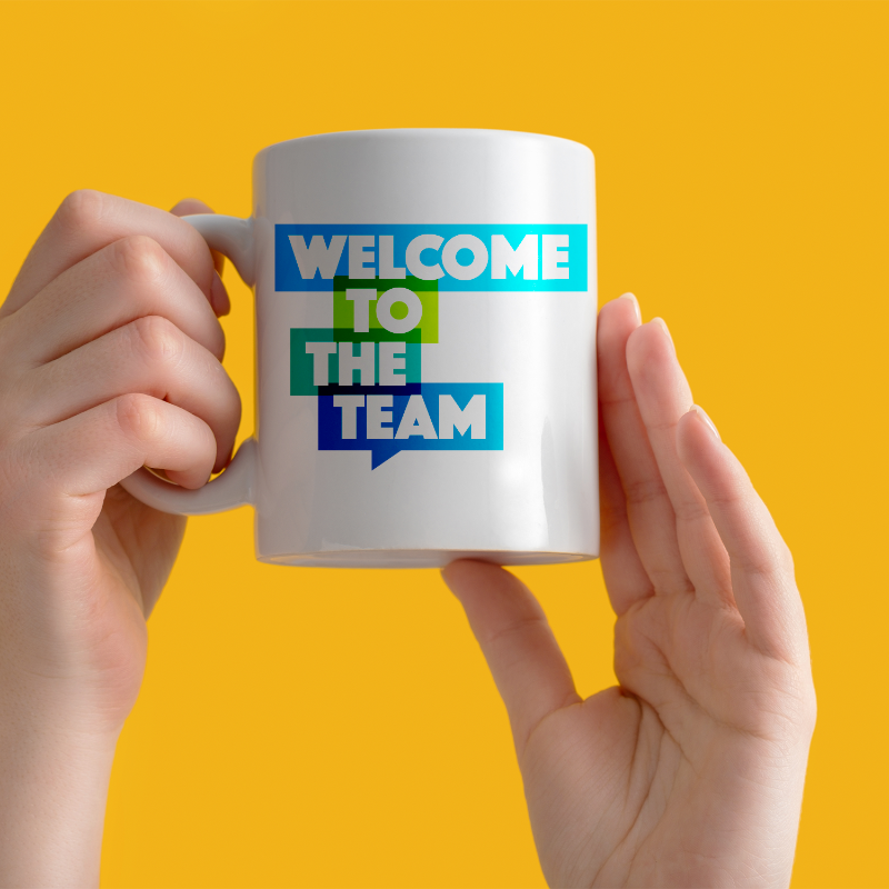 A person holding a white ceramic mug with the text 'Welcome to the TEAM' printed in blue and green colors.