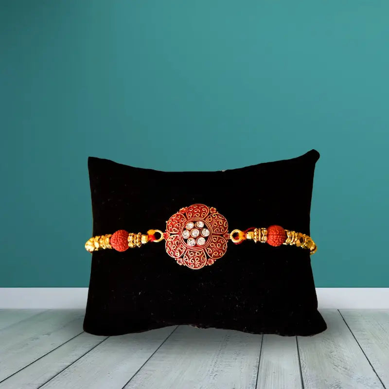 A traditional Indian Rakhi bracelet with a central red and gold filigree medallion, red beads, and gold spacer beads, displayed on a black velvet pillow against a teal wall and light wood floor.