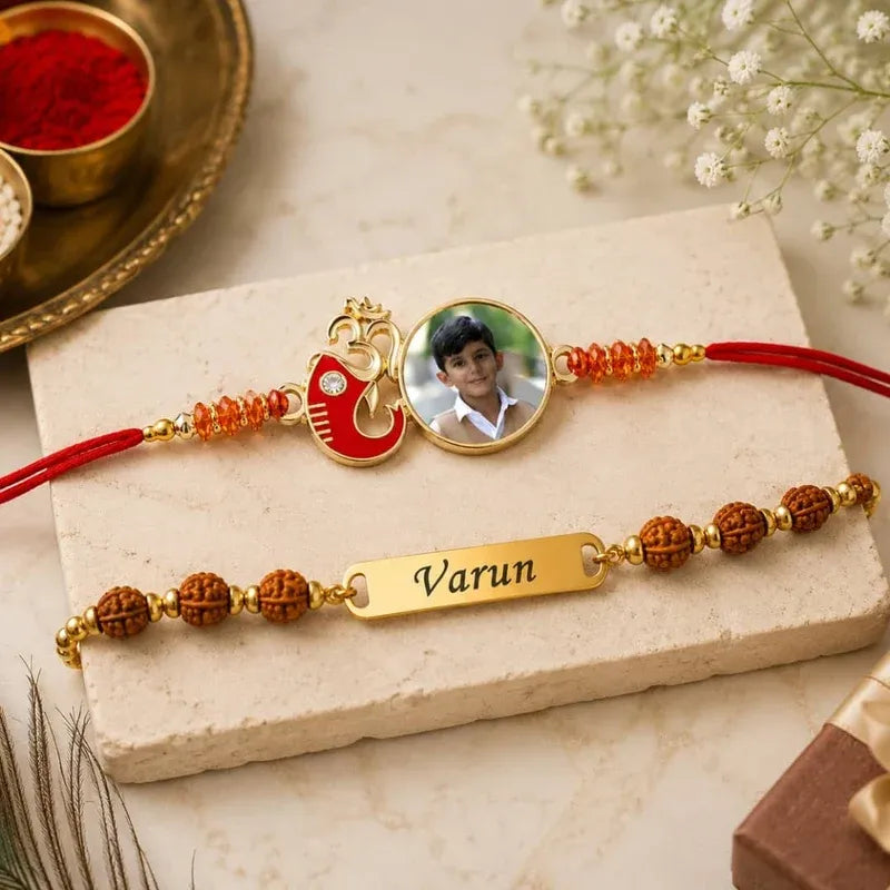 Two custom Rakhi bracelets displayed on a cream stone block. The top Rakhi has a red cord, a red and gold Peacock charm, and a circular photo insert.
