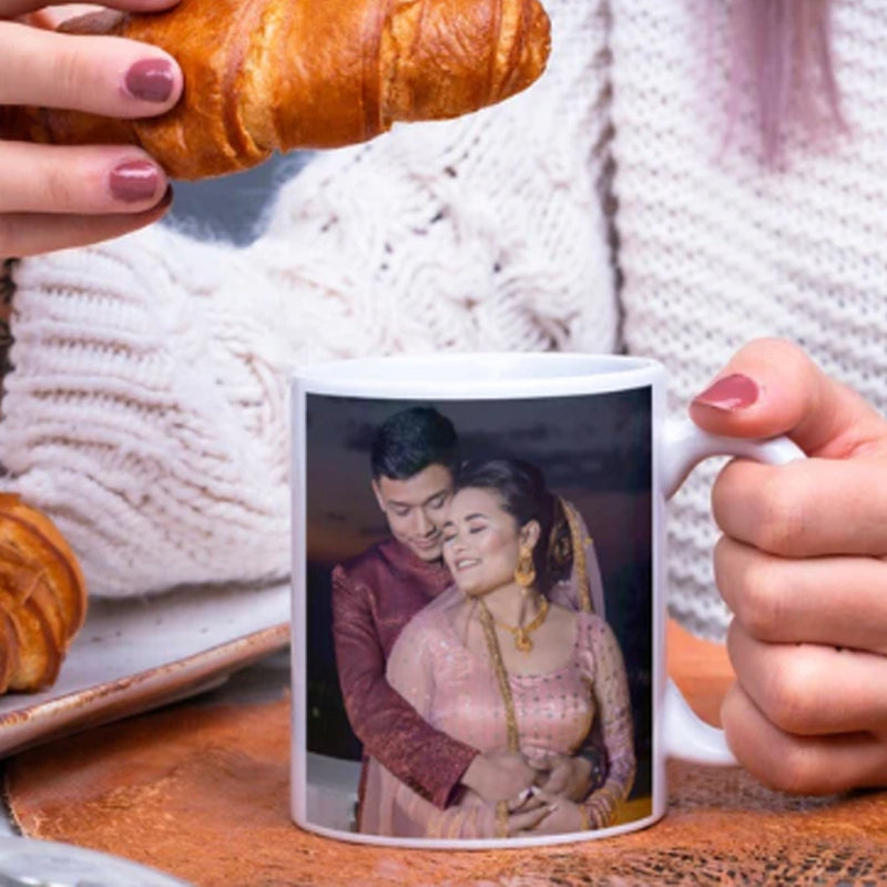 Person holding a mug with a printed image of a couple, next to a plate of croissants.