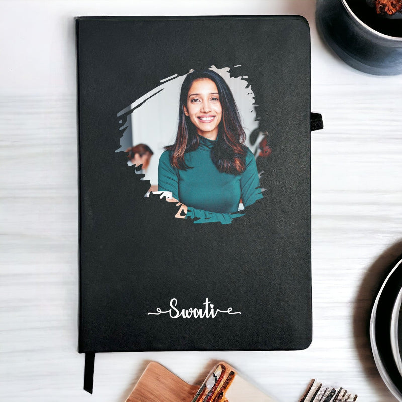 Black notebook with a personalized portrait on the cover, placed on a white surface with a cup of coffee.