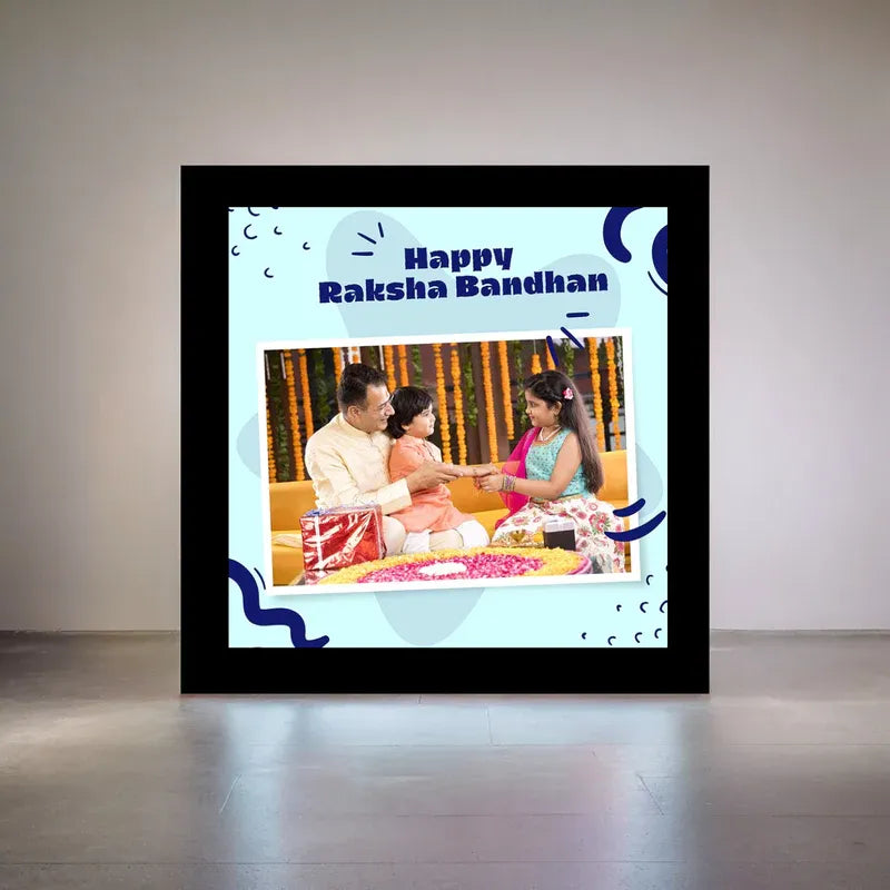 A framed digital photo frame displayed on a wall in a room, showing a custom 'Happy Raksha Bandhan' photo frame design with a black outer border and a light blue inner background. The central photo shows a man, a young girl, and a woman sitting together.