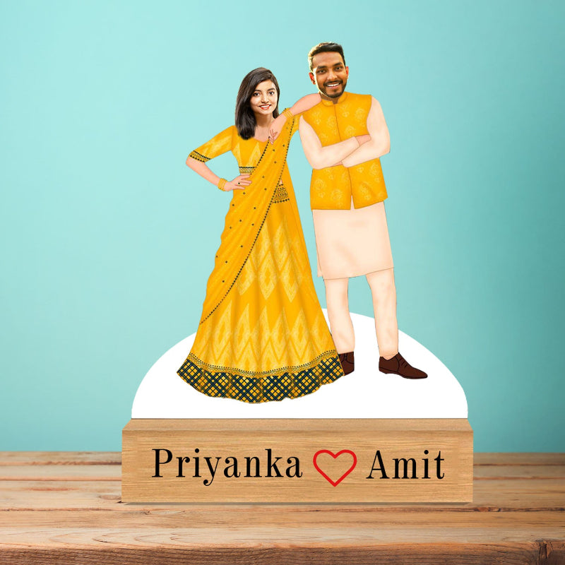 Figurines of a man and woman in traditional yellow outfits on a wooden block with 'Priyanka ❤️ Amit' text against a blue background.