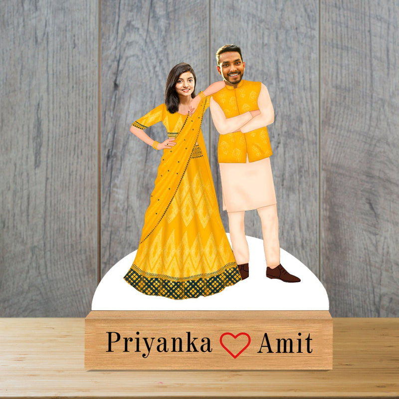 A caricature of a couple, with the man in a yellow suit and the woman in a yellow and green dress, standing on a wooden platform with 'Priyanka & Amit' inscribed at the bottom.
