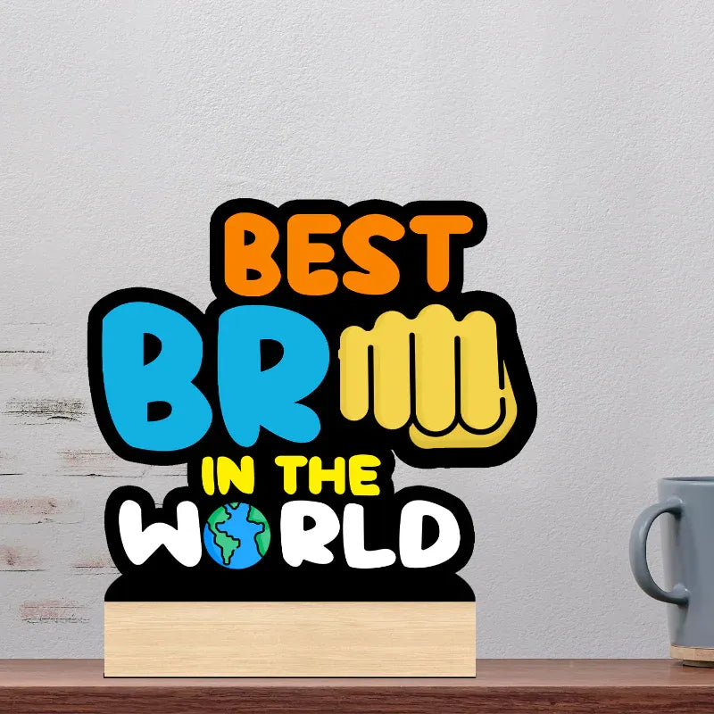 A wooden plaque with a colorful graphic reading 'BEST BR@@M IN THE WORLD' sits on a wooden table against a light gray wall, with part of a gray mug visible on the right.